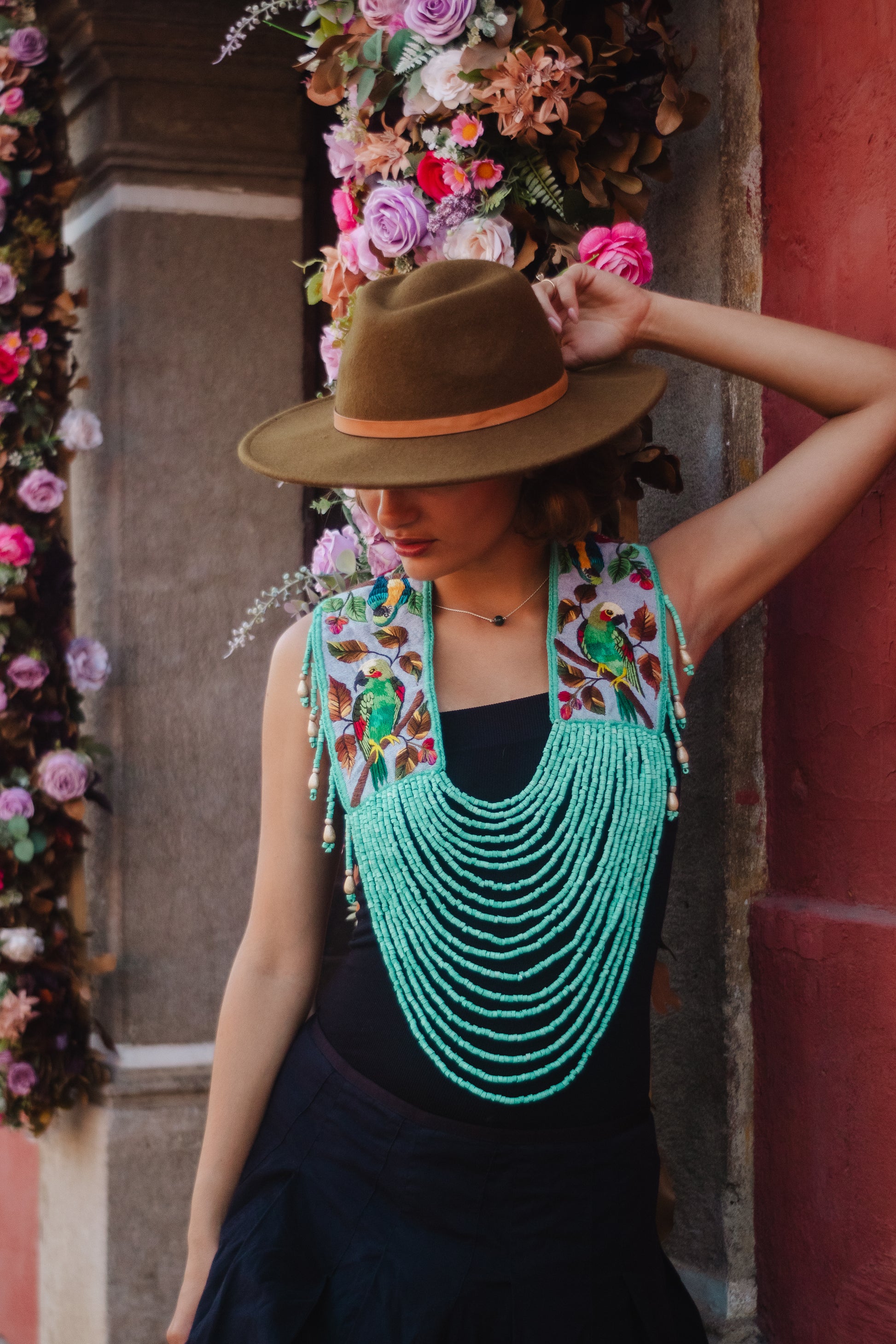 Woman wearing a floral top, green beaded necklace, and brown hat against a decorative wall.