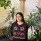 A woman wearing a black long-sleeve dress with a highly detailed, colorful handmade poncho featuring geometric patterns and tassels, posed in front of a warm-toned stone wall and potted green plants.