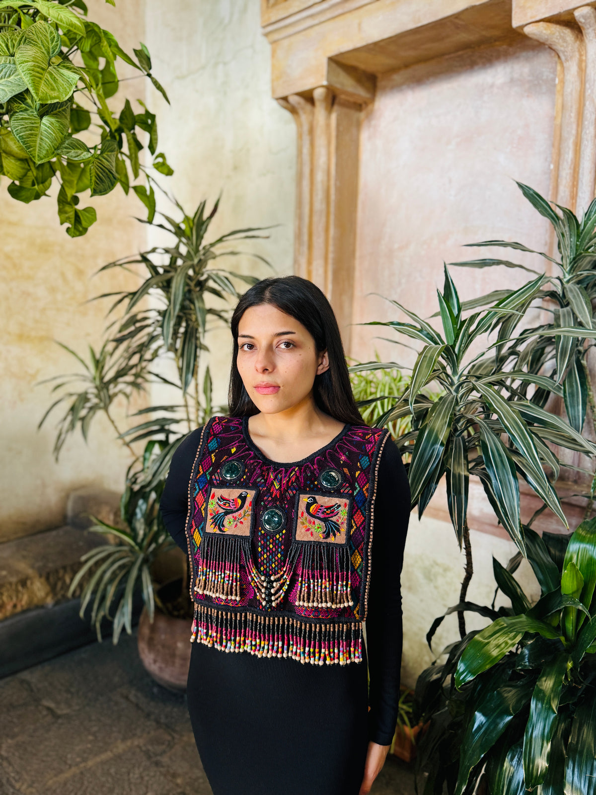 A woman wearing a black long-sleeve dress with a highly detailed, colorful handmade poncho featuring geometric patterns and tassels, posed in front of a warm-toned stone wall and potted green plants.