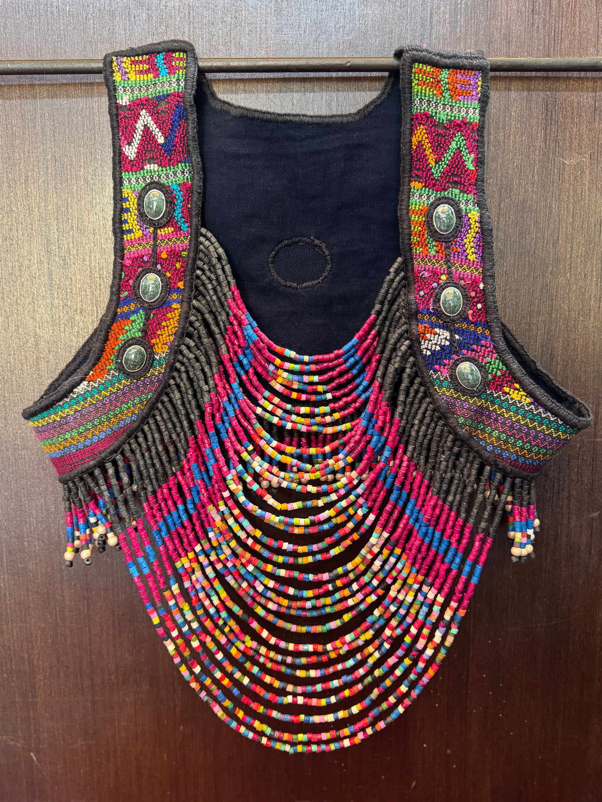 A colorful beaded vest with a dark navy blue fabric base, featuring intricate multicolor beadwork, fringe details, and decorative metal buttons, displayed against a dark wood background.