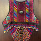 A colorful, hand-woven and beaded vest with intricate geometric patterns in pink, red, blue, yellow, green, and orange, hanging on a dark metal rod against a dark wood background.