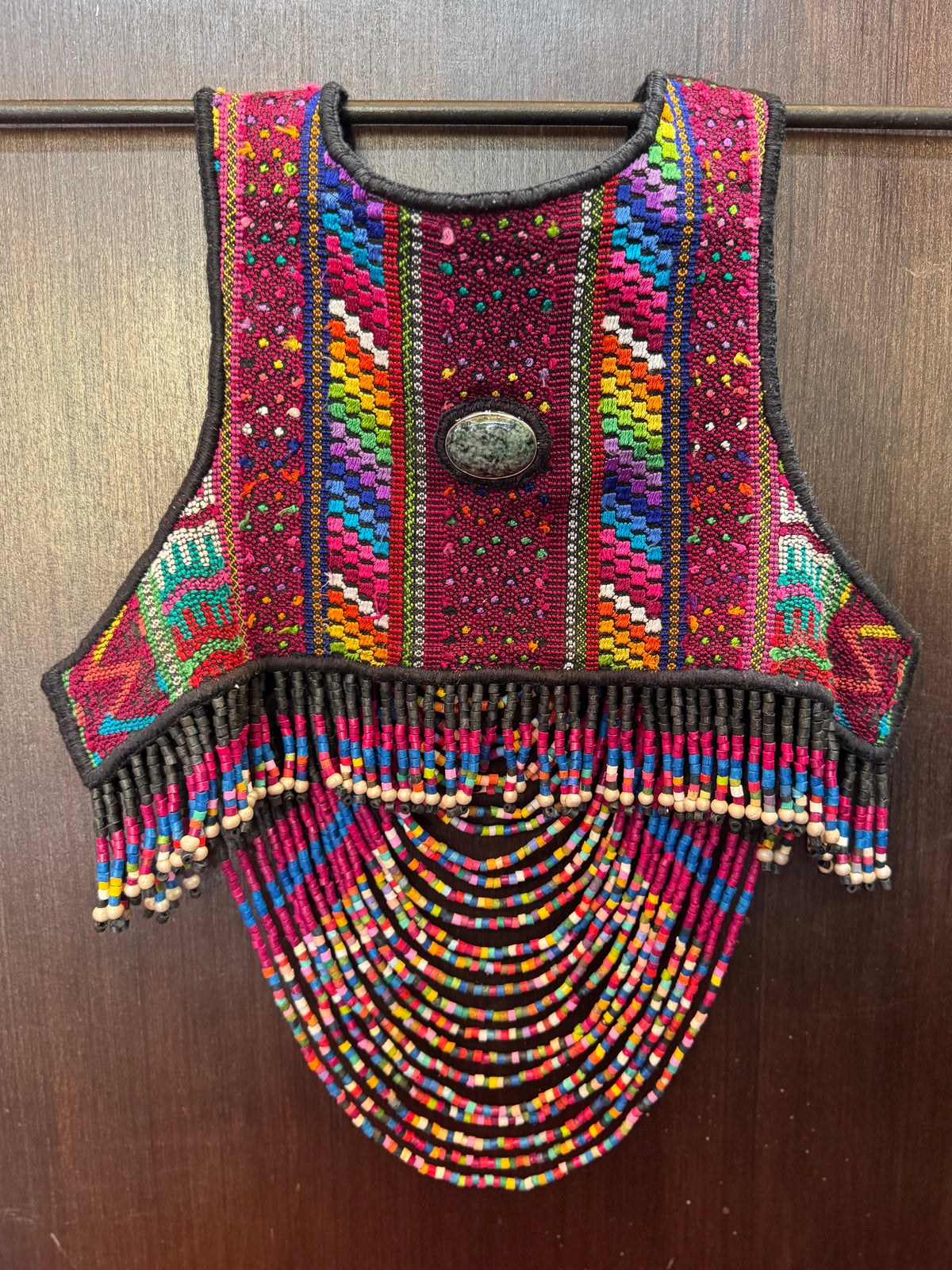 A colorful, hand-woven and beaded vest with intricate geometric patterns in pink, red, blue, yellow, green, and orange, hanging on a dark metal rod against a dark wood background.