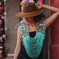 Woman wearing a floral top, green beaded necklace, and brown hat against a decorative wall.