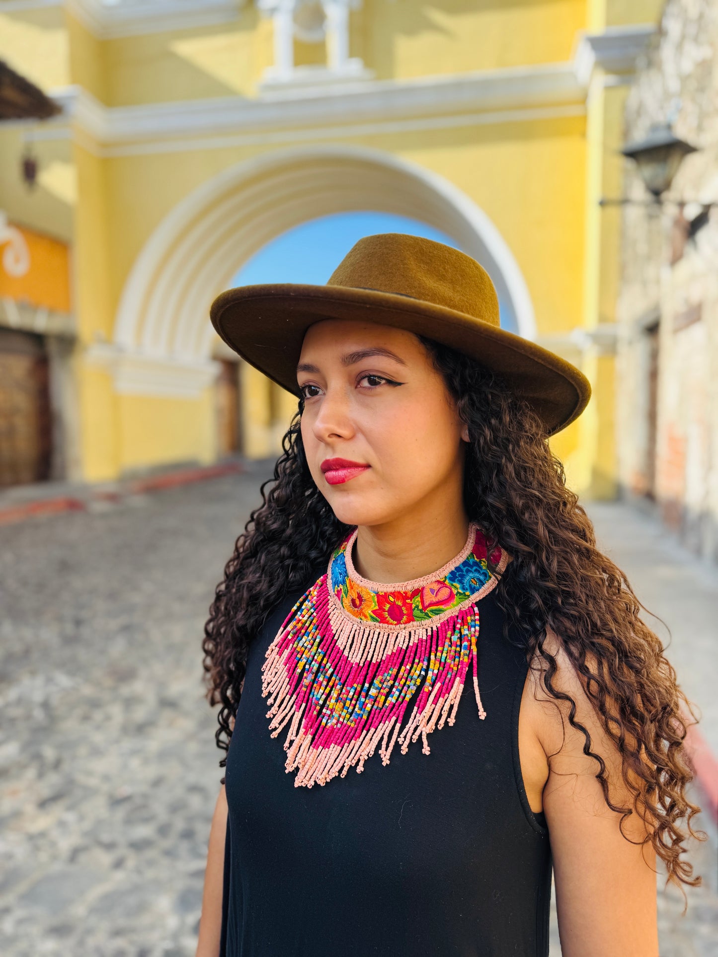 Woman wearing a brown hat and colorful necklace in front of a the famous santa catalina yellow arch in Antigua Guatemala
