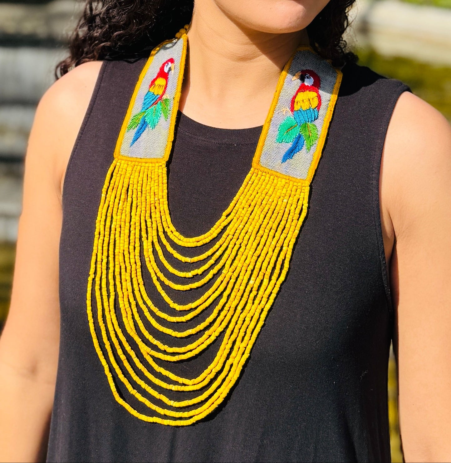 Woman wearing a black sleeveless top with colorful parrot designs and a yellow beaded necklace, outdoors.