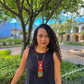Woman wearing a black sleeveless top and colorful necklace outdoors with greenery and buildings in the background