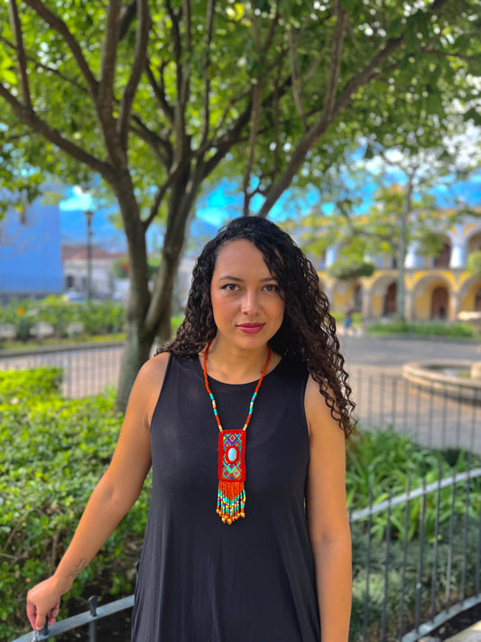 Woman wearing a black sleeveless top and colorful necklace outdoors with greenery and buildings in the background